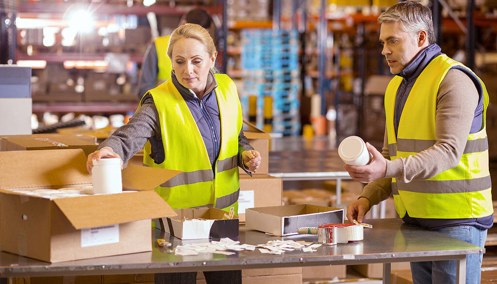 Two Workers In A Distribution Warehouse Placing Products Into A Box Co-Packing For Retail Displays