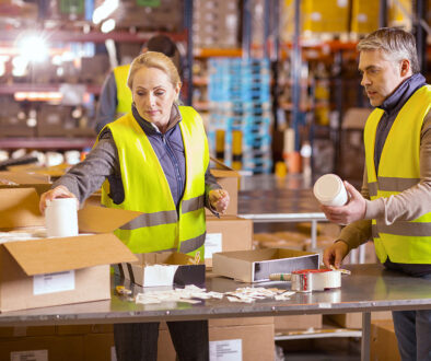 Two Workers In A Distribution Warehouse Placing Products Into A Box Co-Packing For Retail Displays